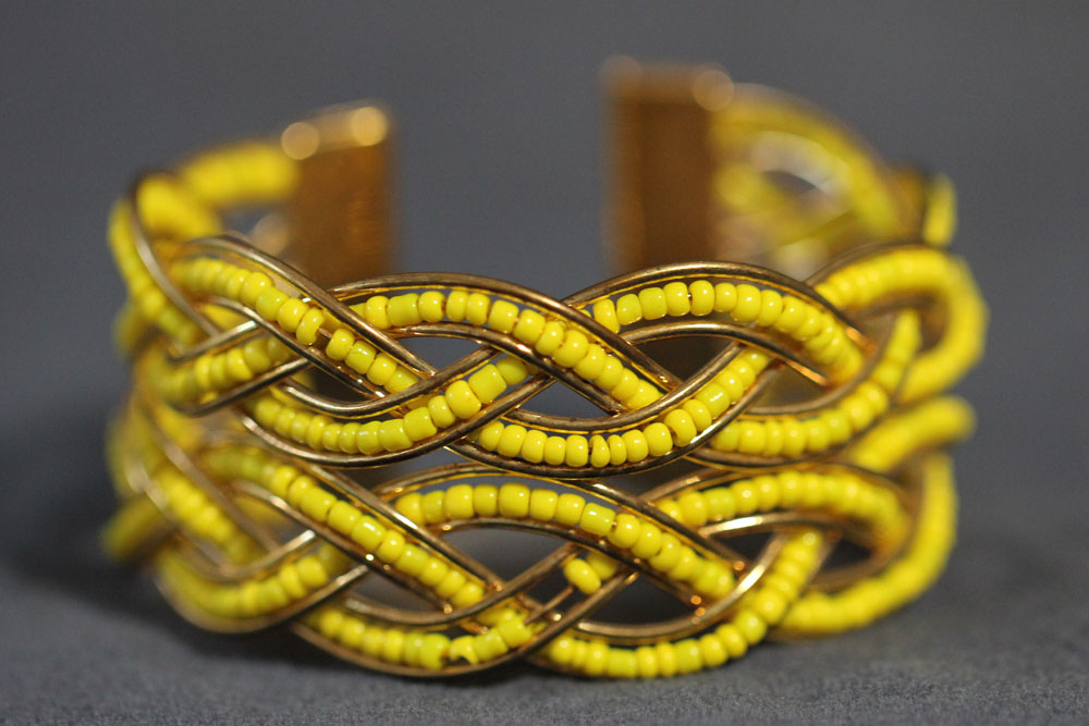 A close-up photo of a wide, gold cuff bracelet with intricate, intertwining designs adorned with many small, bright yellow beads. The background is a muted gray, making the bracelet’s vibrant colors stand out. Consider purchasing this piece as a unique way to support charity.