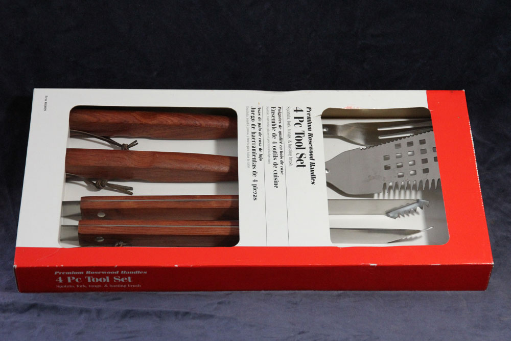 A 4-piece barbecue tool set in a red and white box. The set includes a spatula, fork, and tongs, all with wooden handles. The tools are arranged neatly in the packaging, with product details visible on the box. With every purchase, a portion is donated to charity to provide clean water for those in need.