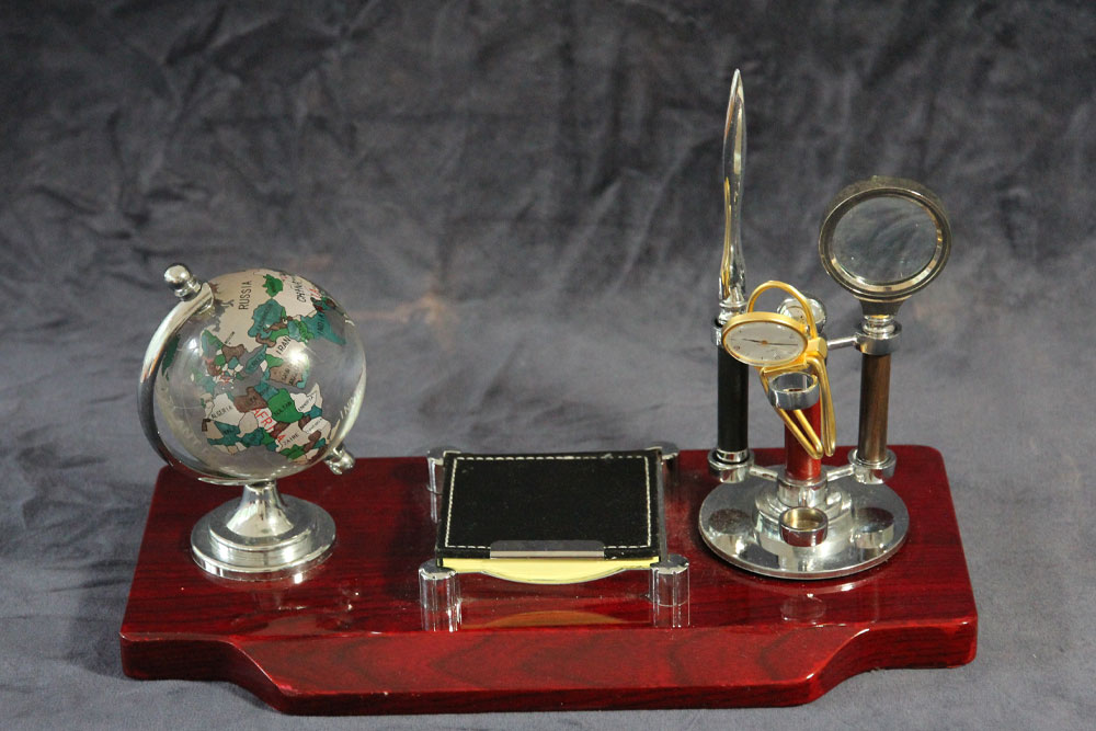 A vintage wooden desktop organizer featuring a small globe, a rectangular notepad, a letter opener, and a magnifying glass. The items are arranged on a polished wooden base with a gray background. Consider making it special by donating yours to charity.