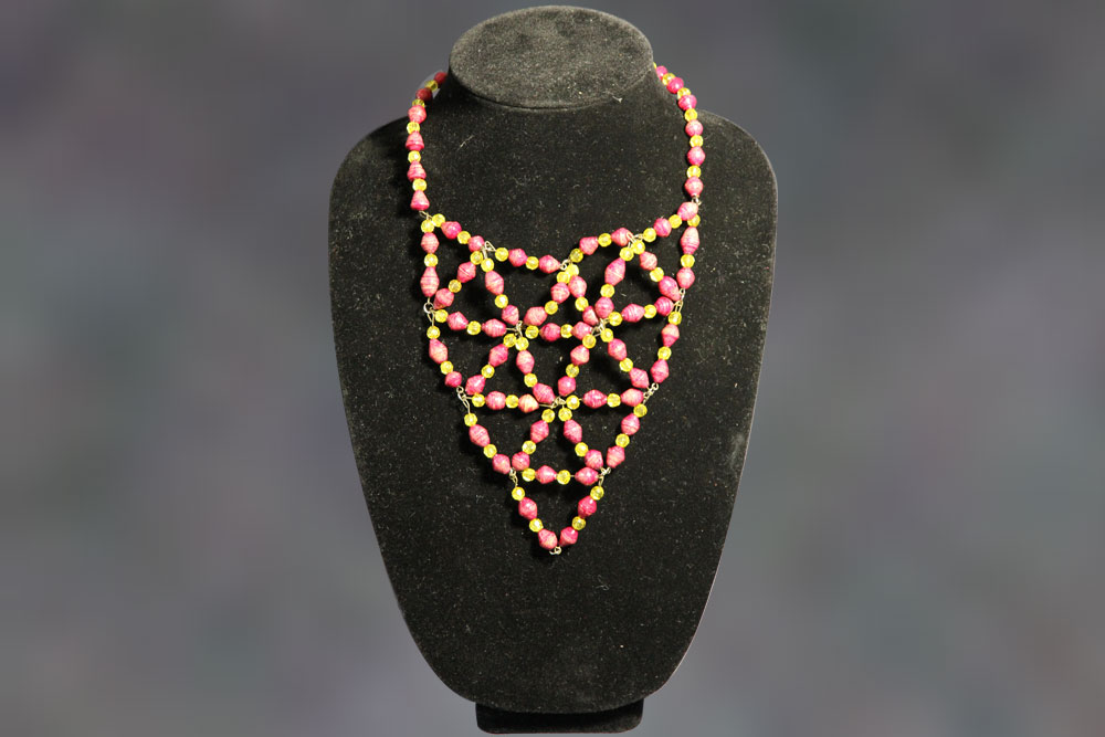 A necklace with a lattice pattern made of pink, gold, and green beads displayed on a black velvet mannequin bust. The necklace forms a triangular design with large diamond-shaped openings. Perfect for charity events, this piece can inspire donations with its elegant craftsmanship.