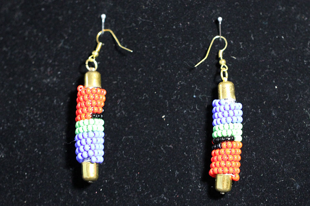 A pair of beaded earrings with gold hooks on a black background. Each earring features cylindrical beadwork in red, purple, green, black, and white, with gold-colored bullet-shaped ends. The intricate pattern celebrates the beauty of diversity while proceeds support water charity donations.