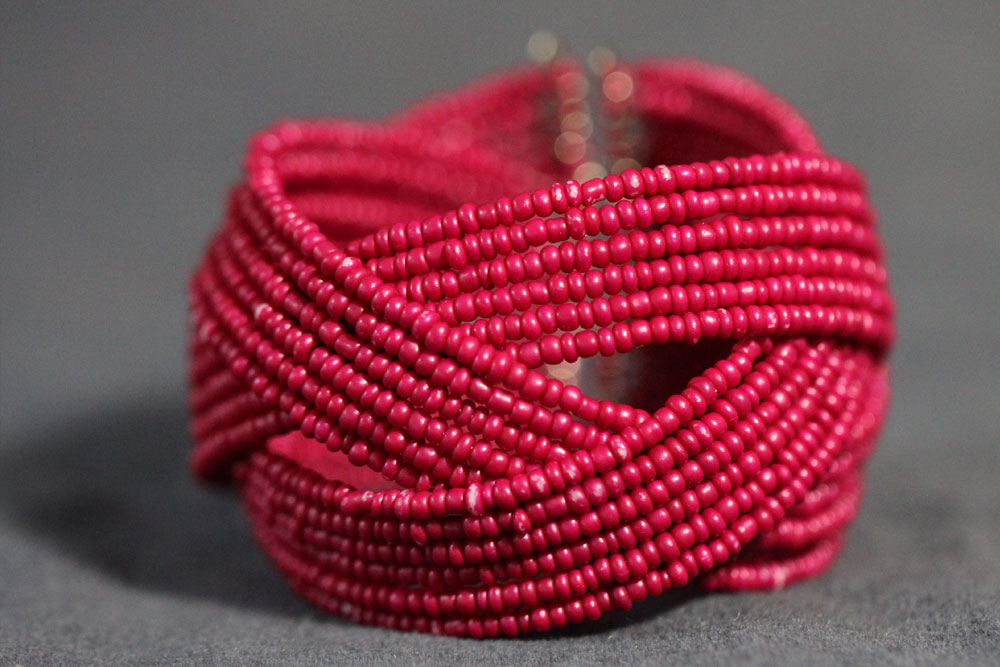 A vibrant pink beaded bracelet, intricately woven in a criss-cross pattern. The bracelet is composed of small, uniformly-sized beads and sits on a smooth grey surface, showcasing its detailed craftsmanship and bright color. By purchasing this piece, you contribute to charity efforts with every donation made.