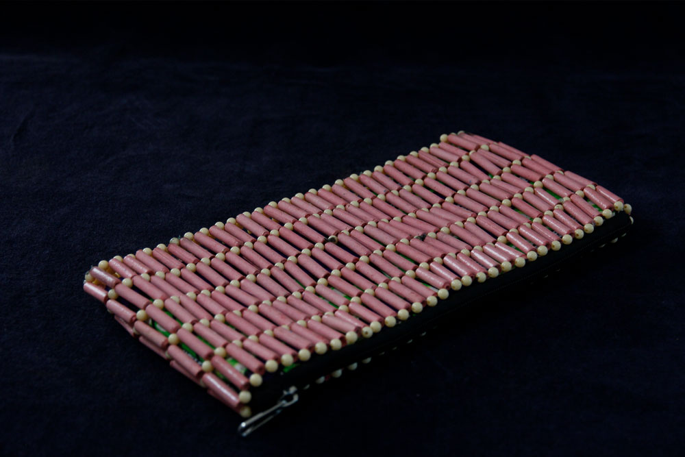 A rectangular, woven clutch on a dark surface. The clutch features rows of cylindrical, pinkish beads interspersed with small, white beads. It has a zipper closure on one of its shorter sides, and purchasing it supports charity efforts through your donation.