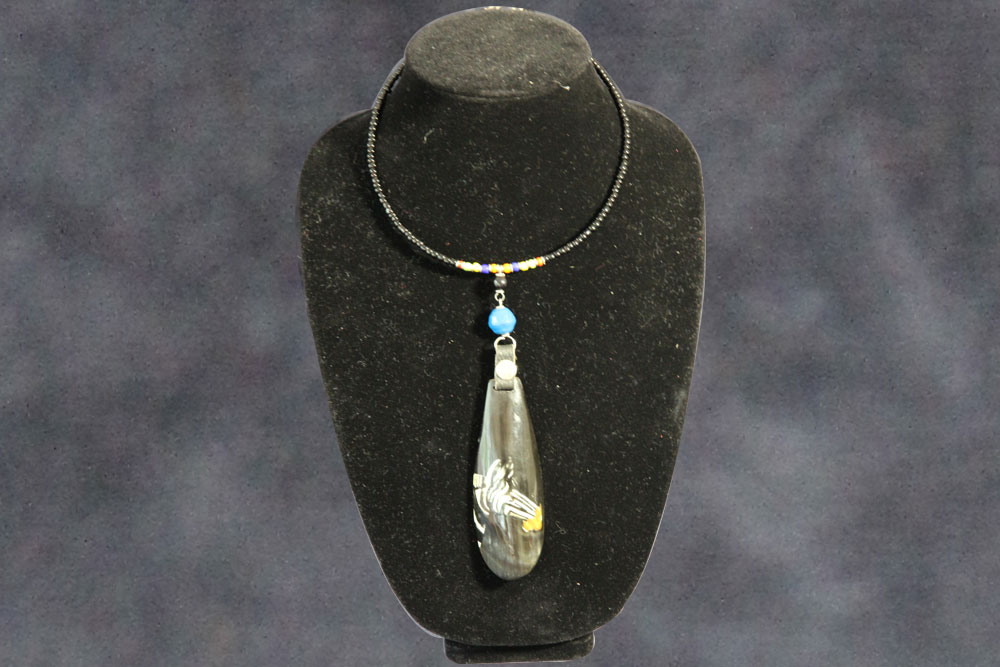 A black beaded necklace with a multi-colored bead accent and an elongated pendant is displayed on a black bust. The pendant features intricate designs and a mix of colors, including a prominent blue bead reminiscent of water near the clasp. The background is a solid, muted color.