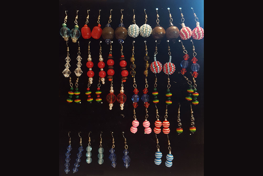 A collection of handmade beaded earrings, created to support charity, displayed on a black background. The colorful earrings are arranged in rows and feature various shapes, colors, and bead combinations, including stripes, solids, and multi-colored patterns. Every purchase helps donate clean water to those in need.