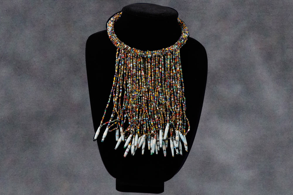 A colorful beaded necklace displayed on a black mannequin. The piece features multiple strands of small, multi-colored beads hanging down in a fringe, each strand ending with white, cylindrical beads. Partial proceeds from the sales are donated to charity, providing clean water to those in need.