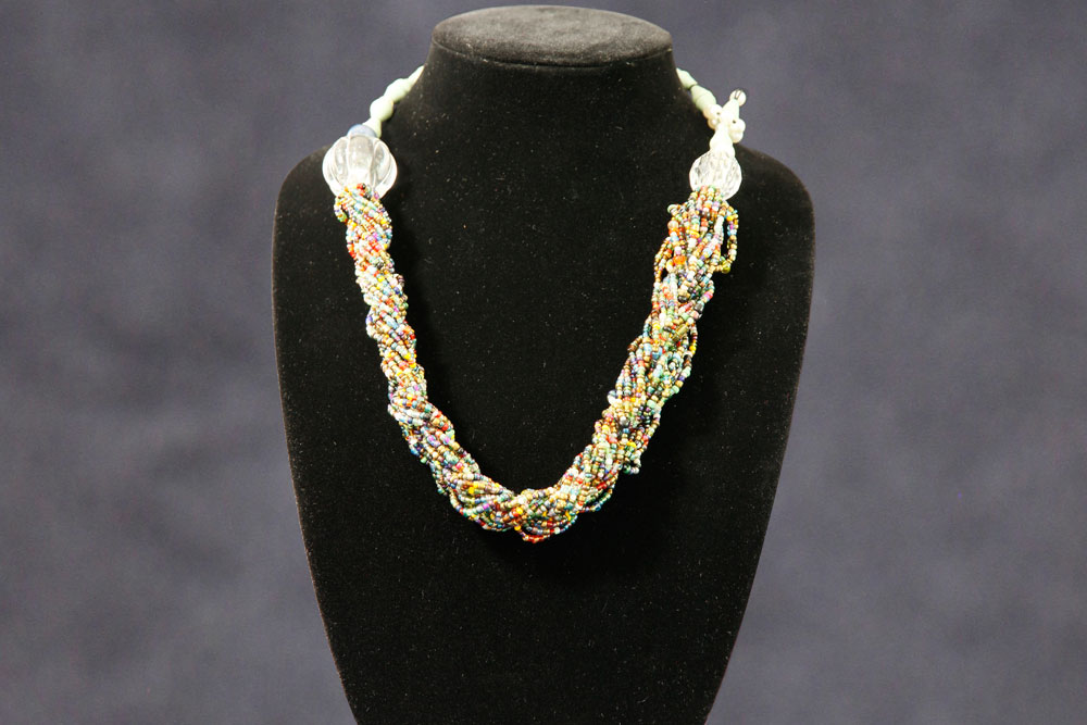 A multicolored beaded necklace displayed on a black velvet bust. The necklace features tightly braided strands of small, colorful beads, creating a vibrant and intricate design. It is set against a dark, plain background, emphasizing the necklace's details. All proceeds from its sale will support charity donations.