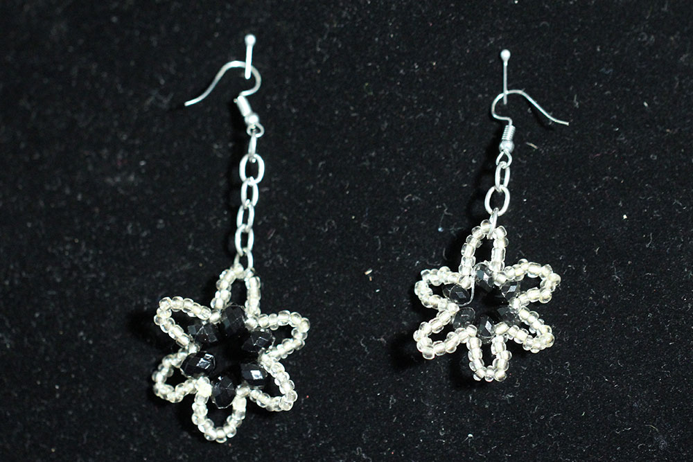 A pair of dangling silver wire earrings on a black background, each designed with a star-shaped pattern adorned with small, shimmering black and clear beads. One earring hangs longer than the other, highlighting a subtle asymmetry. A portion of sales from these earrings goes to water charity donations.