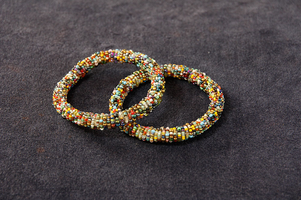 Two colorful beaded bracelets are intertwined and placed on a dark, textured surface. The bracelets, reminiscent of shimmering water droplets, are made up of numerous small beads in various colors, including yellow, blue, red, green, and white, creating a vibrant and intricate pattern.