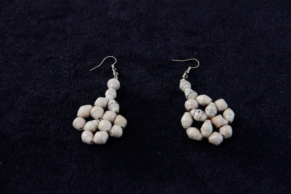 A pair of dangling earrings lies on a dark fabric. Each earring features white beads strung together in a cluster formation, creating a handmade, rustic appearance. The earrings have hook-style closures for wearing. Proceeds from purchases are donated to charity, supporting clean water initiatives.