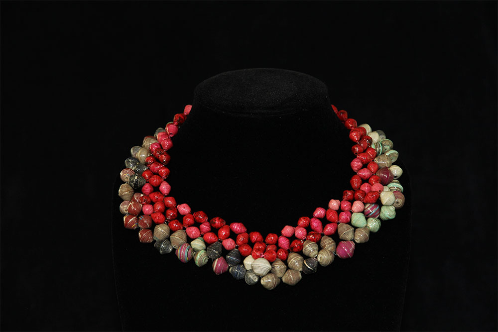 A beaded necklace is displayed on a black mannequin stand. It features clusters of red and green beads in an alternating pattern, forming a layered and vibrant design. The background is black, highlighting the colors of the necklace. Consider this piece for donation to support charity causes.