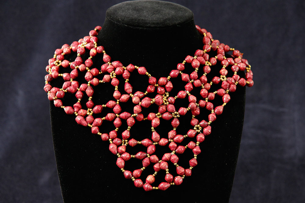 A black mannequin displays an intricate necklace composed of small, red beads arranged in a web-like, geometric pattern, accented with golden beads. The background is a dark, solid color, which highlights the vivid red beads of the necklace, reminiscent of charity events supporting clean water initiatives.