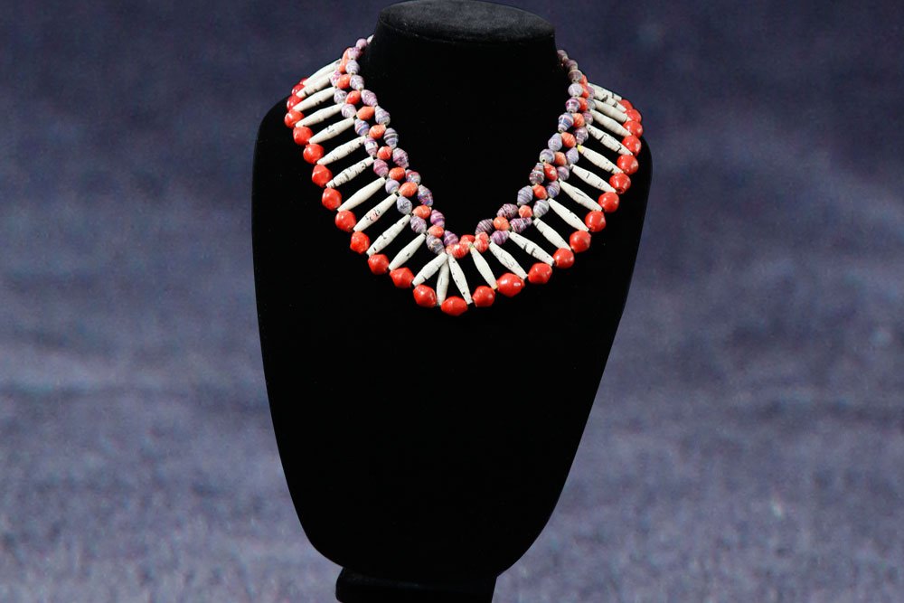 A beaded necklace displayed on a black velvet stand. The necklace features alternating red and white beads with interweaving smaller purple beads, creating a layered and intricate design against a dark blue background. Consider donating to our charity to support more beautiful creations like this.