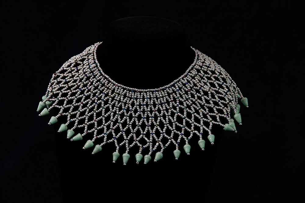 An intricately designed collar necklace made of silver beads, set in a net-like pattern, with green cone-shaped pendants hanging along the edge. The necklace, which was generously donated, is displayed on a black stand against a dark background.