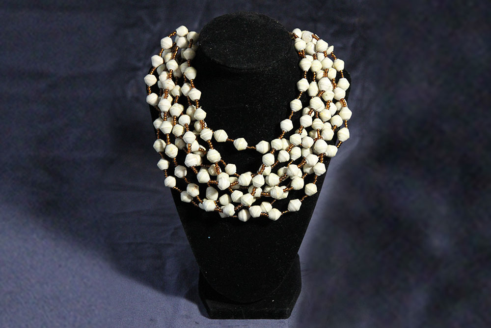 A black display stand holds three necklaces made of white beads with small brown accents. The necklaces are intricately designed with clusters of beads, giving a layered appearance. The backdrop is a dark, textured fabric. Consider donating to charity; each purchase supports clean water initiatives worldwide.