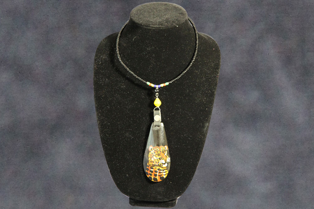 A black necklace with a teardrop-shaped pendant displayed on a black velvet stand. The pendant features an intricately painted leopard face, while the necklace includes small beads near the clasp. A portion of each sale is donated to charity, making this piece both beautiful and impactful. The background is a soft, gradient blue.