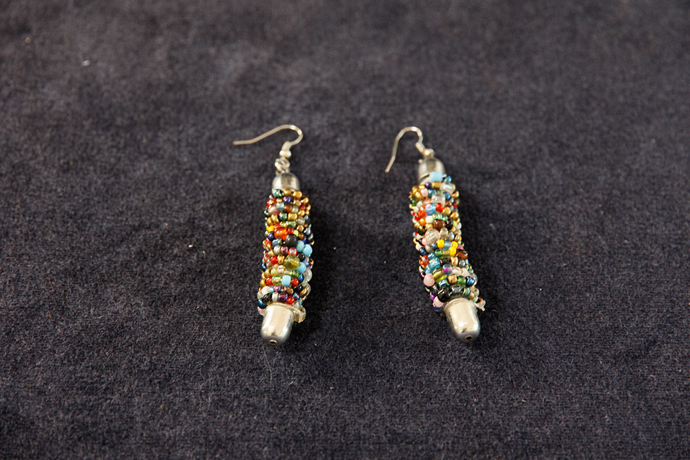 A pair of colorful beaded earrings laid out on a black surface. The earrings feature a mixture of small, multicolored beads intricately woven together around a central, elongated silver base. Each earring has a hook for wearing. Proceeds from your purchase will support water conservation projects via donation.