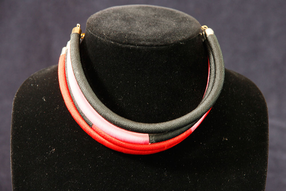 A close-up view of a choker-style necklace displayed on a black bust. The sleek, curved design features segments in black, white, and red, creating a bold, modern look. This stunning piece is showcased against a plain, dark background and exudes an elegance worthy of any charity event.