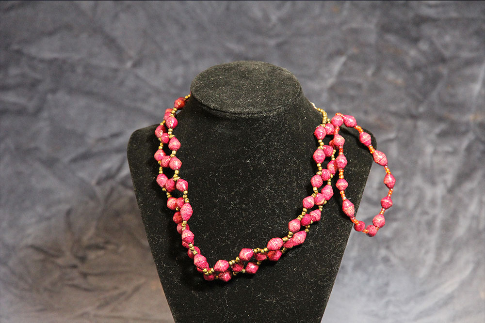 A black velvet necklace display stand showcases a vibrant, double-stranded beaded necklace with matching bracelet. The beads are predominantly pink with small gold accents, creating a striking and elegant ensemble against the dark backdrop, reminiscent of water droplets shimmering in the light.