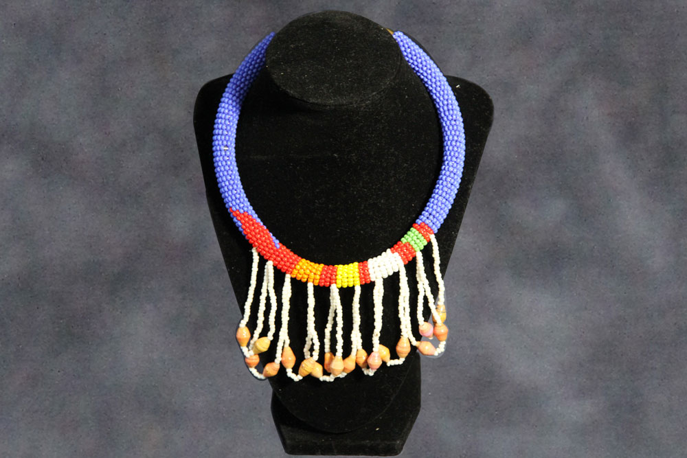 A beaded necklace displayed on a black stand. The necklace is primarily blue with sections of red, yellow, green, and orange beads. White bead strings hang from the bottom, each ending with multiple small beads. The background is a muted, dark gradient. Consider donating to support clean water initiatives.