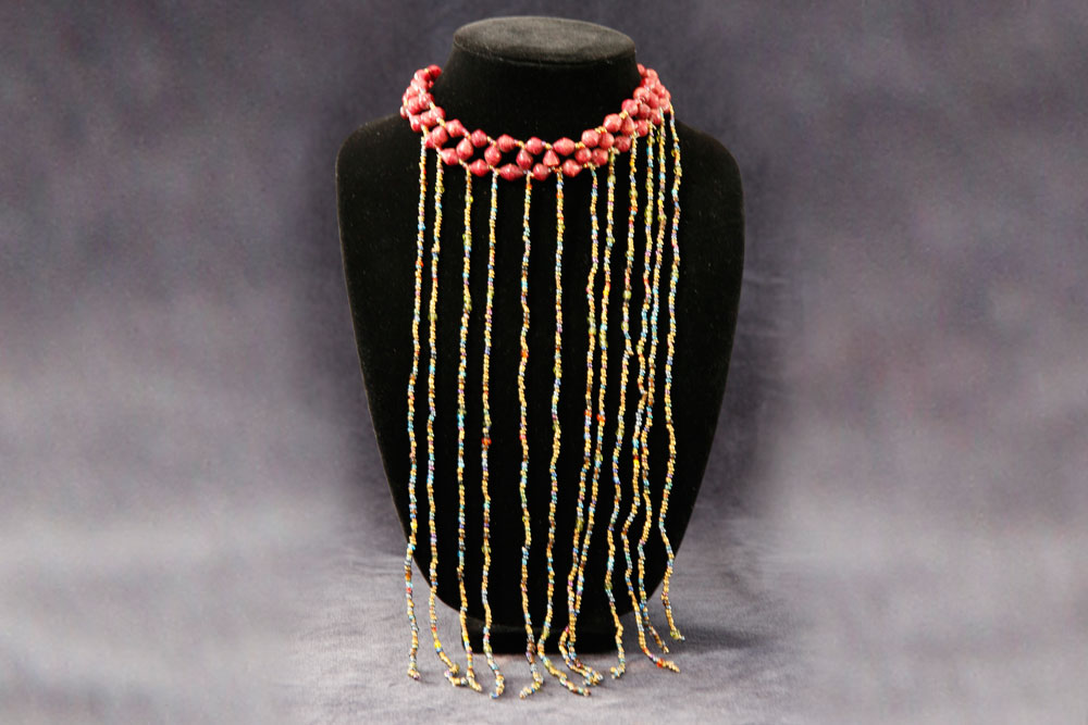 A beaded necklace displayed on a black velvet bust. The necklace features bright red beads around the neck portion and multiple long, dangling strands of multicolored beads in blue, yellow, and red, creating a fringe-like effect that reaches down past the bust. Proceeds from your purchase will help donate clean water to charity.