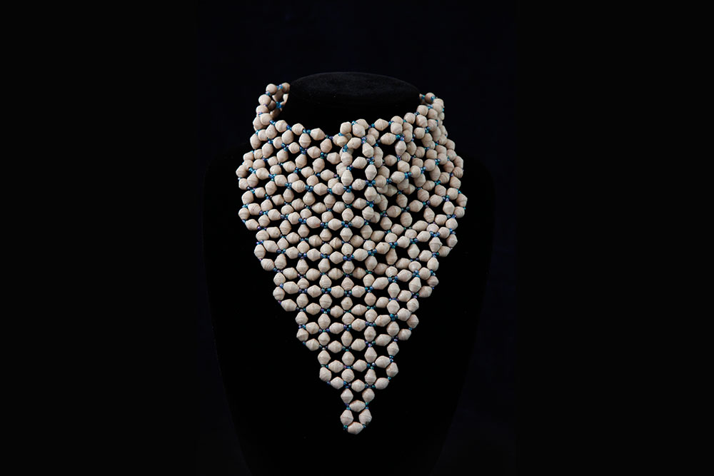 A necklace made of numerous small, spherical, beige-colored beads is displayed on a black bust. The beads, symbolizing purity like water, form a triangular pattern that drapes elegantly downwards. This piece also represents the spirit of charity and donation.