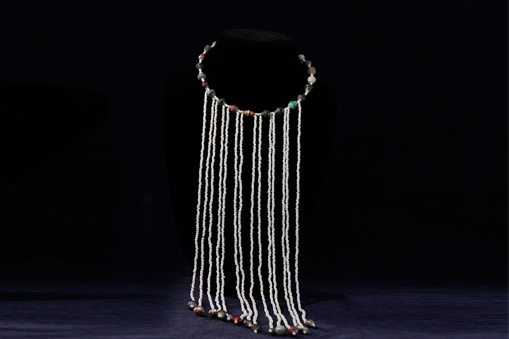 A beaded necklace displayed on a black stand. The necklace consists of variously colored beads and features multiple long strands of white beads that drape down in the front. Consider making a donation to support this charity's cause.