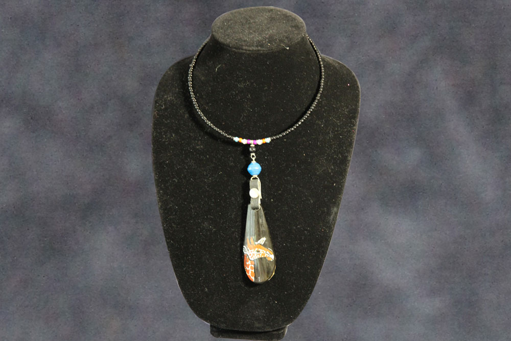 A black beaded necklace is displayed on a black velvet stand. It features a dangling teardrop-shaped pendant with intricate orange and white designs, and a small blue bead symbolizing water above it. The background is a gradient of dark shades, perfect for any charity auction to encourage donations.