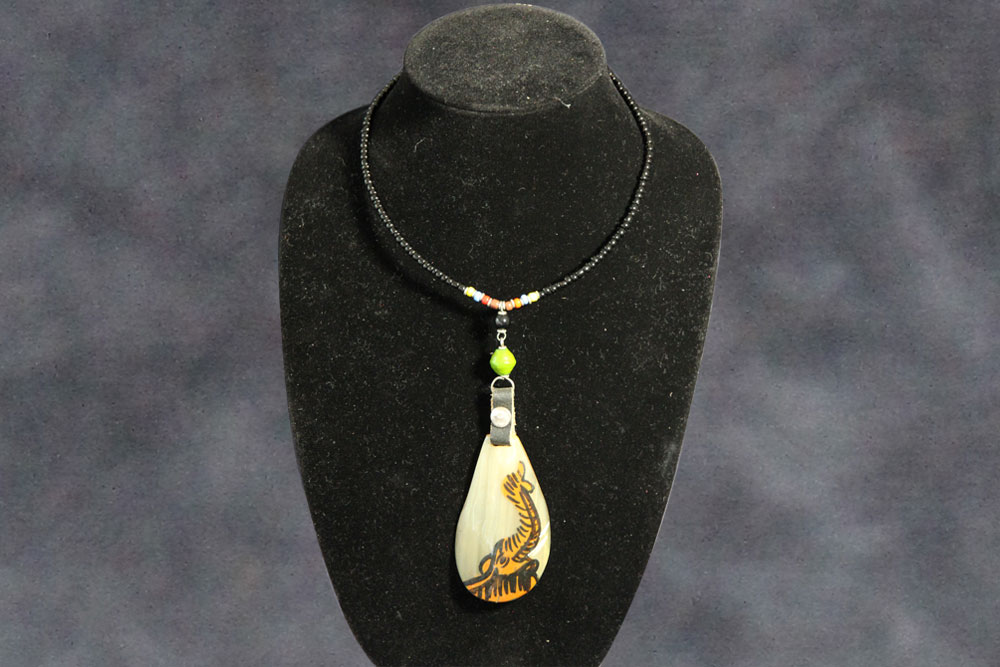 A beaded necklace displayed on a black stand. The necklace features small black beads and decorative elements, including a large pendant with detailed carvings that resemble flora. A portion of the proceeds will be donated to charity for clean water initiatives. The background is a blurred, dark gray tone.