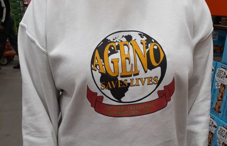 A person wearing a white sweatshirt with an image of a globe and the text "AGENO SAVES LIVES" in large, bold letters. Below it, on a red ribbon, are the words "THE AGENO FOUNDATION INTERNATIONAL, INC." encouraging charity donations to support their noble cause.