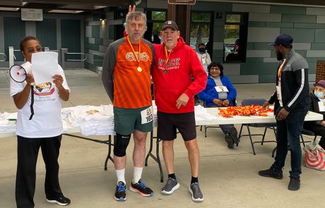 A man in an orange shirt with a medal stands next to a man in a red hoodie, both smiling. The man in the red hoodie has his arm around the other man. Nearby, a person holds a megaphone and a paper. Several people are seated at a table with shirts, medals, and donation jars for charity.