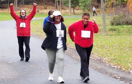 Four people are participating in an outdoor charity race on a paved park trail surrounded by autumn trees. Two women in the front are cheering, one in a white cap and the other in a red hoodie. They are followed by a man in a red jacket with his arms raised and another participant.