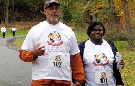 Two individuals dressed in white event t-shirts, one labeled with the number 102 and the other with 101, walk hand-in-hand on a paved path in a park with autumn foliage. Both appear to be participating in a 5K walk/run event, with donation stations offering water to participants along the way.