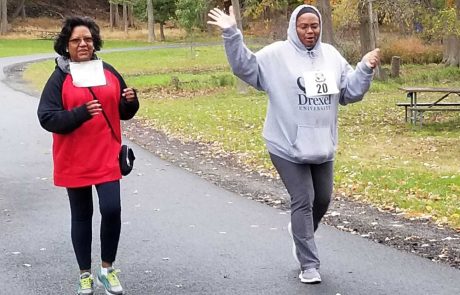 Two women are walking on a paved path in a park surrounded by autumn foliage. One wears a red and black jacket, black pants, and glasses, the other sports a grey Drexel University hoodie and grey pants, with arms raised in a cheerful manner. They seem to be participating in a walk event supporting water donation initiatives.

