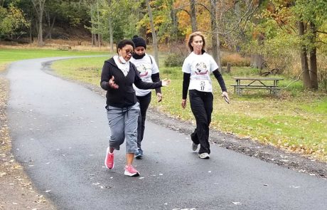 Three people are walking on a paved path in a park, surrounded by autumn trees. Two women, wearing matching white T-shirts and black pants, are walking together, while another woman in a black jacket and gray pants walks slightly ahead with bright pink shoes. One of the women hands out water to passing joggers as a gesture of donation.