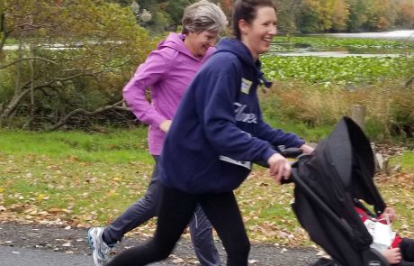 Two women jog along a paved path near a lake surrounded by autumn foliage. One woman, wearing a navy hoodie and pushing a stroller, smiles as they pass the shimmering water. The other, in a pink jacket, jogs slightly behind and to the side. Both appear to be enjoying the run.