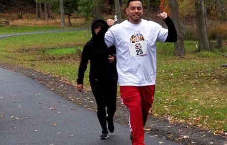 Two people are jogging on a paved path in a park surrounded by trees. The person in front, wearing a white T-shirt with the number 25 and red pants, is smiling and flexing both arms. The person behind, dressed in black with a hood and face partially covered, is holding a small sign that reads "Donate to Charity.