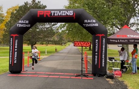 A runner in a white shirt and black pants crosses the finish line under a black PR Timing inflatable arch with a red digital timer displaying 2:26:02. Two people stand near the timing tent on the right, observing the charity event. Trees and a path are in the background.