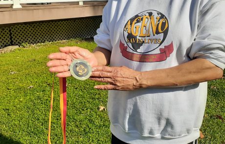 A person wearing a white sweatshirt with a "SAGENO SAVES LIVES" logo is holding a medal. The medal has a ribbon in red and yellow colors. The background shows a grassy area and part of a wooden structure, symbolizing the success of their charity initiatives encouraging people to donate for clean water projects.