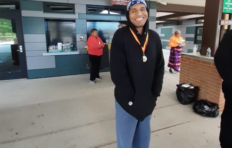 A smiling person is standing outdoors wearing a black hoodie, blue sweatpants, and a headband, proudly displaying a medal around their neck. Three other people are in the background near a building with a snack stand and water station. One person is holding a clipboard for the charity event.