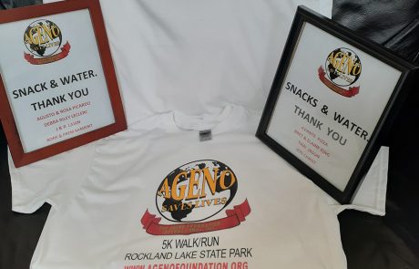 A display featuring a white t-shirt with the logo "AGENO Saves Lives" and event details for a 5K Walk/Run at Rockland Lake State Park. Framed signs reading "Snack & Water Thank You" with donor names are placed on either side of the t-shirt, highlighting generous donations.