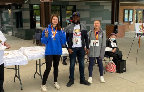 Three people in athletic attire pose for a photo at an outdoor event. They are wearing numbered bibs and medals around their necks. Behind them, a table with event t-shirts and donation boxes is visible. Most people are wearing casual clothes, holding water bottles, and the atmosphere appears festive.