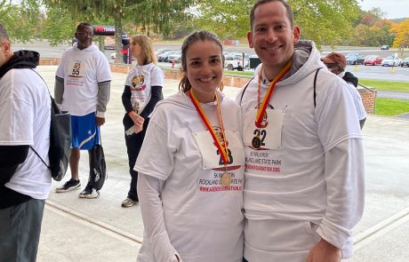 A smiling man and woman wearing white event t-shirts and medals stand close together, posing for a photo outdoors. Other participants with similar attire and race bibs are in the background, preparing for or finishing a race. Trees, parked cars, and a water station for donations are visible.