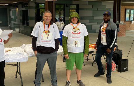 Three men wearing race medals and event t-shirts pose for a photo at what appears to be a race event registration or finish area. They stand under a pavilion with tables and additional t-shirts in the background, where participants are encouraged to donate to charity while staying hydrated with provided water.