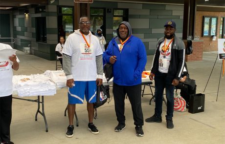 Three men wearing race numbers and medals stand together under a shelter at an outdoor charity event. The man on the left is in a white t-shirt and shorts, the middle man in a blue hoodie, and the right man in a black jacket and cap. A table with donation materials is beside them.