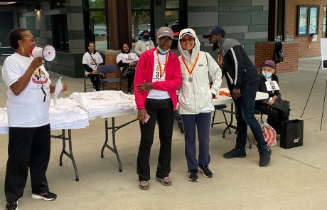 A group of people at an outdoor event. Two individuals in the center wear medals and white jackets, smiling for the camera. A person on the left holds a megaphone near a table with folded white shirts and a donation box. Others in the background sit or stand near the event setup, enjoying the day.