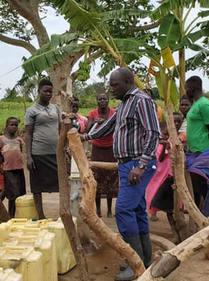 A man, dressed in a striped shirt and blue pants, operates a water pump surrounded by several people, including children and adults. In this outdoor setting with trees and jerry cans on the ground for collecting water, their efforts highlight the importance of charity to support access to clean water.