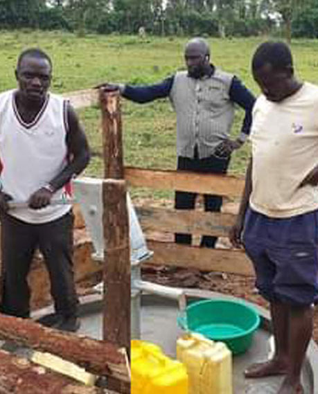 Three men are gathered around a hand pump water well in a rural setting. One man is actively pumping water, while the other two watch. A yellow jerry can and a green bucket are positioned near the well to collect precious water. Green fields are visible in the background, showcasing the importance of donation efforts for such essential resources.