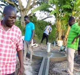 Four men are standing near a recently constructed well. One man, in a checked shirt, stands to the left while two others, in blue and green shirts, stand by the well. The fourth man works in the background. Thanks to a generous donation, lush greenery surrounds the scene.