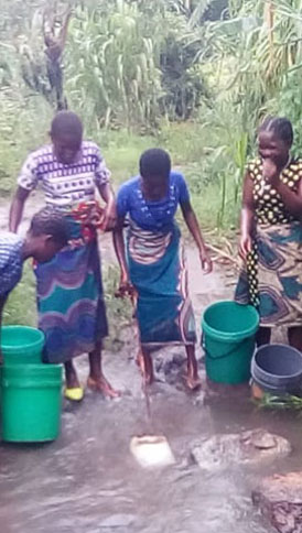 Three people are standing in a shallow stream, using buckets to gather water. The individuals are wearing colorful clothing and are surrounded by lush green vegetation. One person leans over to fill a bucket, while the others watch and assist. Their efforts highlight the importance of water conservation and encourage donations for clean water initiatives.